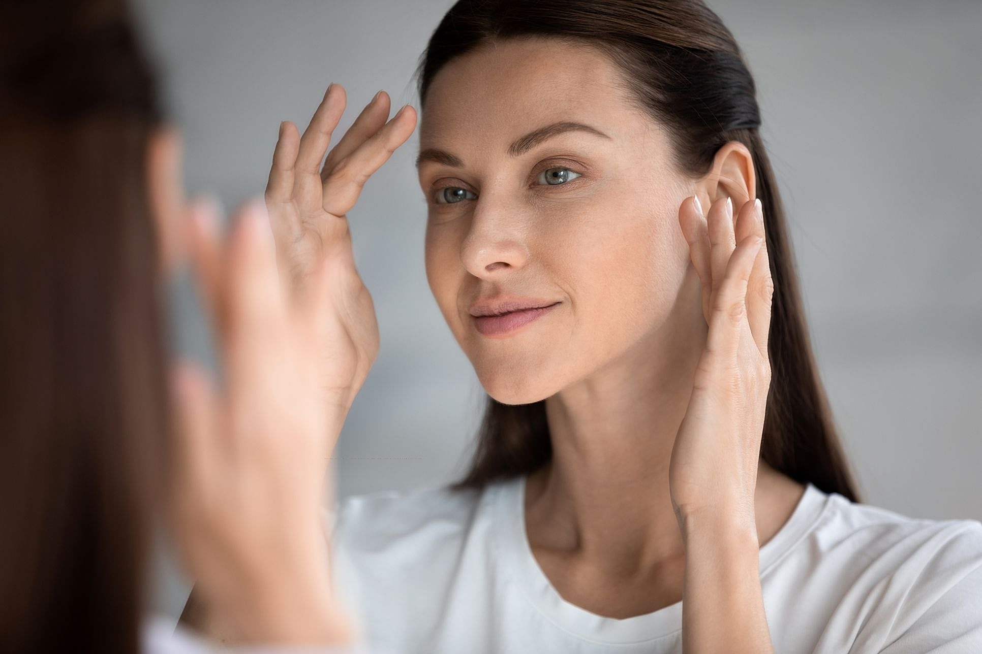 A woman gently touching her face in the mirror during her facelift recovery journey to check for swelling and healing progress.